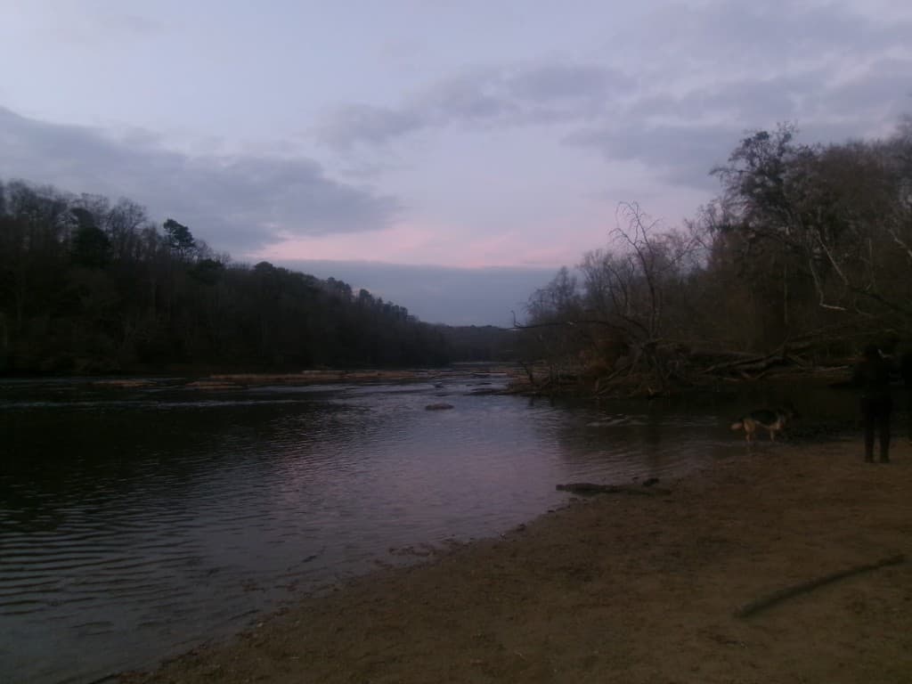 River at dusk with forested banks, a person and dog on the shore